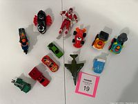 Top-down view of 12 assorted toy vehicles and action figure spread out on flat white surface including cars, a plane, a boat, and a red action figure.