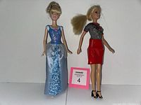 Front view of two Barbie dolls, one in a blue and silver formal gown and the other in a casual black and red outfit, posed standing on a white background with a pink lot number card nearby.
