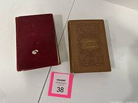 Photo showing two old books placed side by side on a white surface; red cloth bound book with worn edges and brown cloth bound book with embossed decorative front cover.