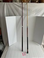 Two full-length wooden pool cues standing upright with a white backdrop, showing leather grips, metal rings, and decorative ends.