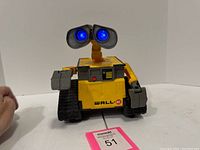 Front view of yellow WALL-E robot toy with lighted blue eyes and articulated arms, sitting on a white surface with a pink label.