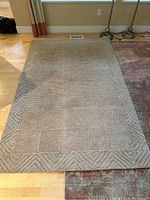 Front view of the hand tufted wool area rug showing the textured geometric squares and border pattern in neutral beige tones laid on hardwood floor next to another rug.