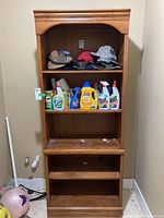 Wooden bookcase in laundry room, showing three shelves with detergent bottles and hats on top.