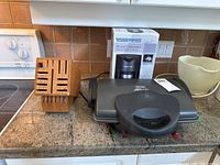 Overall view of the coffee maker box, wooden knife block and healthy grill on kitchen counter.