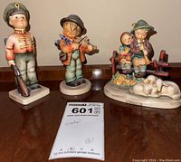 View of all three Goebel porcelain figurines; two boys and one girl with musical instruments and animals, arranged on a table.