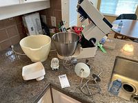 Photo showing Kenwood Chef stand mixer with stainless steel and plastic bowls plus multiple attachments on countertop.