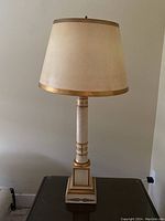 Tall table lamp unlit showing beige shade with gold trim and gold-accented white column and base, photographed from the front.