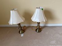 Two brass base table lamps with off-white bell-shaped fabric shades, both lamps placed side by side on carpeted floor against wall.