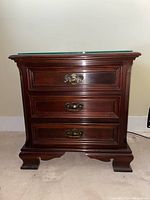 Front view of mahogany nightstand showing three drawers, brass handles, and glass top.