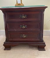 Front view of the solid mahogany nightstand showing three drawers with ornate brass handles and removable glass top, revealing scratches on the surface and drawer fronts.