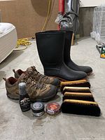 Black rubber boots and brown hiking boots alongside three shoe care brushes and three shoe care product containers on garage floor.
