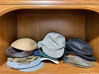 Overall view of multiple men's hats displayed on a wooden shelf, showing various colors and materials, mainly wool flat caps with one leather cap.