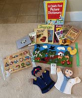 Overview photo showing all items in the lot on floor: A Child's First Picture Dictionary, other small children's books, two felt hand puppets, two wooden puzzles (animal and shapes), small wooden toy.