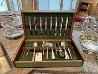 Silverplate flatware in green felt-lined wooden box, showing knives, forks, spoons arranged in slots.