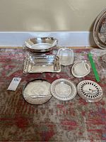 Photo showing various silverplate serving trays and bowls arranged on a carpet, showcasing different shapes including oval, rectangular, and round.