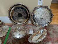 Photo showing four silverplate serving pieces against a beige wall and red patterned rug including two large trays, one small bowl, and one oval tray with ornate designs and edges