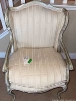 Front view of upholstered occasional chair showing beige striped fabric, wooden carved frame with distressed white finish, and removable seat cushion.
