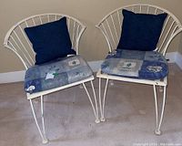 Set of 2 white wrought iron chairs with floral seat cushions and blue pillows, showing overall condition and slight paint chipping.