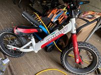White and red Infinity balance bike with black tires and no pedals shown in a cluttered environment.