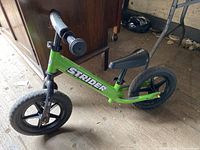Green Strider balance bike with black wheels, black seat and handle grips placed indoors.
