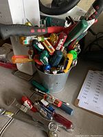 Photo showing a metal bucket filled with various screwdrivers, wrench tools, and a hammer, some tools placed on the surface outside the bucket, with a printout of numbers partially visible.
