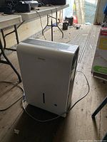 Photo of a white Danby dehumidifier unit placed on a wooden floor near table legs with power cord plugged in and visible.