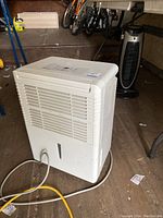 White Danby dehumidifier visible from front-left side in a dim indoor space with bikes and a standing fan visible in the background. Unit plugged in with white cord.
