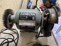 Photo showing the Delta 6-inch bench grinder with wire brush wheel on the left side, grinding wheel on the right, motor housing with product labels, power cord, and tool rest.