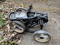 Folded black three-wheeled golf push cart showing two larger rear wheels and smaller front wheel with mesh basket area and straps.