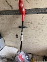 Full view of Homelite electric trimmer showing red handle, black shaft, protective guard and cutting head at bottom wrapped in plastic.