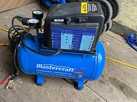 Side view of a blue Mastercraft air compressor with black control unit and attached pressure gauges, showing brand logo, labels, and control switch.