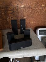 Five Panasonic surround sound speakers: four small vertical satellite speakers and one horizontal center speaker, placed on a surface against a wood background.
