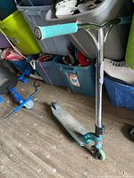 Photo of teal kick scooter with chrome handlebar and teal grips on wooden floor surrounded by storage bins and miscellaneous items.