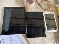 Three tablets placed side-by-side on a textured wood surface: Apple iPad, Huawei, and Samsung; screens are off and reflective with smudges visible.