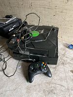 Two black original Xbox consoles stacked, showing green Xbox logos, with one black wired Xbox controller in front and AV cables on top.