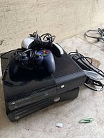 Photo shows two black Xbox 360 consoles stacked, with two black wired controllers and one power adapter cable resting on top, arranged on a textured surface.