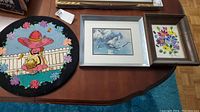 Three framed art pieces on a wooden table with the round textile rug visible next to them.