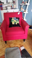 Front view of red upholstered chair with black floral pillow on seat. Shows chair arms, cushion, and pillow detail.