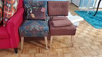 Two occasional chairs side by side on a wooden parquet floor, one brown fabric with pillows and one blue floral fabric with a floral pillow.