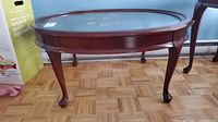 Oval wooden coffee table shown from one angle, showing the beveled clear glass inset top and one polished wood cabriole leg with claw foot.
