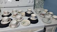 Large assortment of teacups and saucers arranged on stove showing various floral and gold detail patterns.
