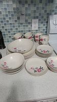 Photo showing cups, saucers, dessert bowls, side plates, bowls, and dinner plates with pink rose floral design arranged on kitchen counter.