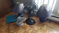 Overview of the lot showing two table lamps, two fans (one black Holmes desktop, one smaller white fan), and a blue scale on parquet floor.