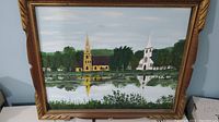 Framed original painting showing churches of Mahone Bay with reflection on water and surrounding trees. Painted in oil or acrylic on canvas or board.