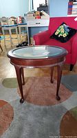 Oval wooden side table with beveled glass top and carved cabriole legs, shown from a slight angle with red armchair and other furniture in background.
