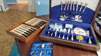 Wood table with silverplate flatware sets including a wooden box with knives and forks, and a blue velvet case with multiple silver utensils.