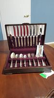 Wooden canteen box with silverplate flatware neatly arranged including knives, forks, spoons and a small spreading knife. Box is lined with burgundy velvet and satin.