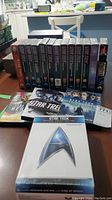 Photo showing twelve Star Trek VHS tapes lined up with colorful covers and titles, plus four Star Trek DVDs including the Original Motion Picture Collection in a box set case.
