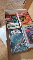 Photo of an open box containing multiple Star Trek paperback novels with several visible book covers including one by William Shatner and 'Star Trek: The New Voyages' featuring a starship on the cover.