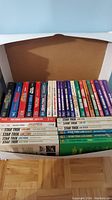 Two boxes filled with Star Trek novels showing colorful spines with titles and authors, arranged horizontally in two rows inside open boxes on a wooden floor.